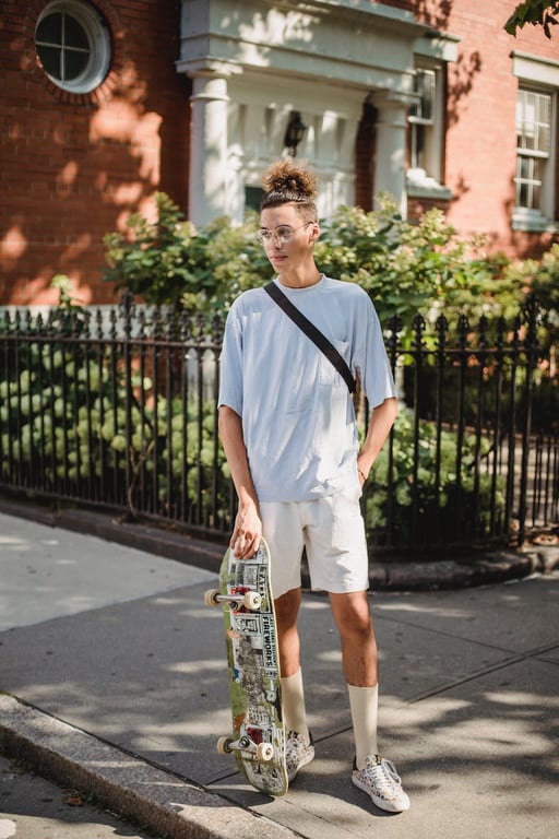 Corps entier d'un jeune homme ethnique aux cheveux bouclés, portant des vêtements élégants et des lunettes, se reposant sur le trottoir près d'un bâtiment en briques avec un skateboard et regardant au loin.